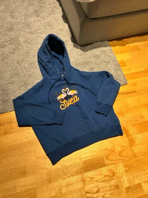Blå hoodie från Svea med flamingos - Snygg blå hoodie från Svea med tryck av två flamingos och texten 'Svea' i gult framtill. Hoodien har huva med dragsko och ribbade muddar vid ärmslut och nederkant. 👍Perfekt för en avslappnad och cool stil.🙌🏻 Perfekt för en chill dag!
