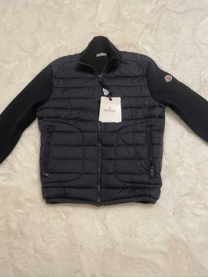 Svart Cardigan från Moncler - Snygg svart cardigan från Moncler med quiltad front och stickade ärmar. Jackan har hög krage, två dragkedjeförsedda fickor och Moncler-logga på ena ärmen. Perfekt för kalla dagar och ger en stilren look.