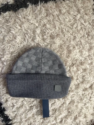 Grå Petit Damier nya Säsong - Säljer en grå Petit Damier  med klassiskt rutmönster och ribbad uppvikt kant. Mössan har en diskret LV-logga på en liten patch framtill. Tillverkad i mjuk ullmix som håller dig varm och snygg under kalla dagar.