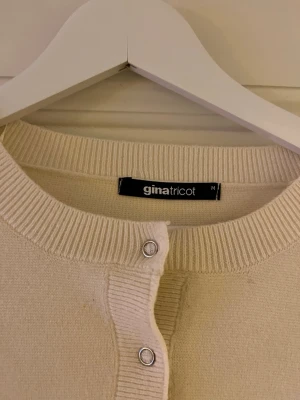 Beige kofta från Gina Tricot - En stilren beige kofta från Gina Tricot i storlek M. Koftan har rund halsringning, ribbade muddar och knappar framtill i silverfärg. Materialet är mjukt och stickat, perfekt för lager-på-lager och en clean look.