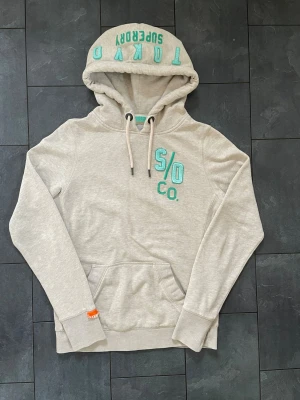 Beige/grå hoodie från Superdry Tokyo - Snygg beige/grå hoodie från Superdry Tokyo med turkosa broderade detaljer på bröstet och insidan av huvan. Klassisk känguruficka framtill, dragsko i huvan och ribbade muddar. Perfekt för en avslappnad streetwear-look. St S.