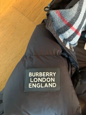 Svart pufferjacka från Burberry - Snygg svart pufferjacka från Burberry med stor logotyp på ärmen och klassiskt rutigt foder i huvan. Jackan har dragkedja, knappar och två fickor framtill. Perfekt för kalla dagar och riktigt trendig look.