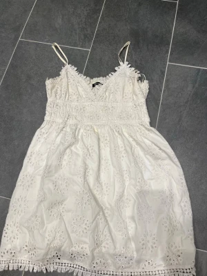 Vit spetsklänning med broderi från Ginatricot - Söt vit kortklänning med tunna axelband och v-ringad hals. Klänningen har broderade blommönster och spetsdetaljer längs kanterna samt en smockad midja för skön passform. Perfekt för sommarens alla tillfällen!