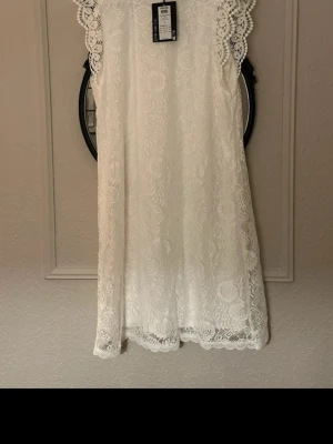 Vit spets klänning från Pieces - Söt vit klänning från Pieces i spets med fina virkade detaljer vid axlarna. Blusen är ärmlös och har rund halsringning. Perfekt till sommaren, student eller skolavslutning 