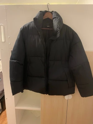 Svart pufferjacka från BikBok - Säljer en svart pufferjacka från BikBok i recycled material. Jackan har hög krage, dragkedja framtill och två stora fickor på sidorna. Perfekt för kalla dagar och ger en riktigt snygg streetwear-look.