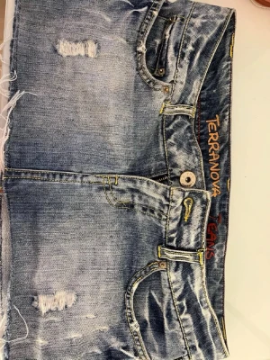 Blå jeanskjol från Terranova - Säljer en blå jeanskjol från Terranova med slitna detaljer och rå kant nertill. Kjolen har klassisk femficksdesign, knapp och dragkedja framtill samt gul kontrastsöm. Perfekt för dig som gillar en avslappnad och trendig stil.
