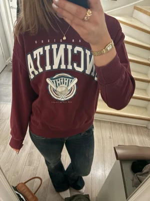 Vinröd sweatshirt med tryck - Säljer en vinröd sweatshirt med stort vitt och blått tryck på bröstet och ett basebollmotiv. Tröjan har rund hals, ribbade muddar och är i mjukt material. Perfekt för dig som gillar sportig och avslappnad stil.