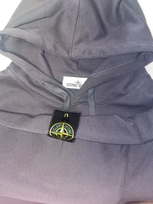 Mörkgrå hoodie från Stone Island - Snygg mörkgrå hoodie från Stone Island med klassisk logga på ärmen. Hoodien har huva med dragsko och en stor magficka framtill. Tillverkad i mjukt bomullsmaterial som känns skönt mot huden. Perfekt för en avslappnad och trendig look.