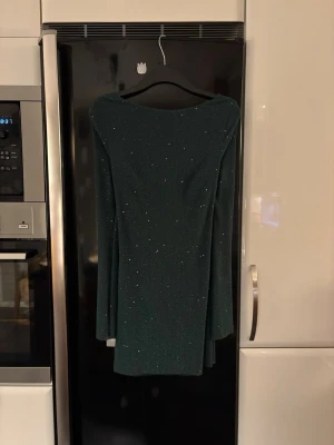 Mörkgrön glittrig klänning med öppen rygg - Snygg mörkgrön kortklänning med glittrigt tyg och långärmad design. Klänningen har en djup öppen rygg med draperad effekt och knytband i nacken. Perfekt för dig som vill sticka ut med stil och elegans.