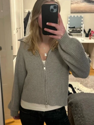 Grå ribbstickad zip-kofta - Säljer en grå ribbstickad kofta med dragkedja framtill och hög krage. Koftan har långa ärmar och är gjord i ett mjukt material som känns skönt mot huden. Perfekt att slänga på sig när det är lite kyligt.