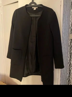 Svart enkel kappa från H&M - Stilren svart kappa från H&M med rak passform och öppen front. Kappan har två stora fickor framtill och är helfodrad med slätt tyg. Perfekt för dig som gillar minimalistisk och klassisk stil. storlek 40 men är mer som 38