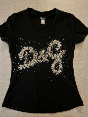 Svart D&G t-shirt med paljetter - ett stort D&G-logo framtill dekorerat med glittriga paljetter. T-shirten har en rund halsringning och är tillverkad i ett mjukt material som sitter tight och markerar figuren. Vintage
