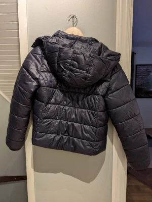 Svart pufferjacka från Gina Tricot - Snygg svart pufferjacka från Gina Tricot med huva och quiltad design. Jackan har en kortare passform och breda kanaler som ger en modern look. Perfekt för kalla dagar när du vill hålla dig varm och samtidigt se trendig ut.