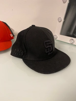 Svart SD keps från New Era - Säljer en svart keps från New Era med broderad SD-logga framtill och initialerna MG på sidan. Klassisk flat brim och strukturerad passform. Kepsen är tillverkad i ett slitstarkt tyg, perfekt för dig som gillar streetwear och sportig stil.