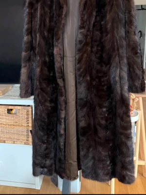 Mörkbrun lång pälsjacka - Säljer en lång, mörkbrun minkpäls i klassisk modell med slät insida i brunt tyg. Jackan har en rak passform och bred krage, perfekt för kalla vinterdagar. Pälsen är tät, mjuk och glansig vilket ger en exklusiv och elegant känsla.   Mått: 	Ärmlängd: 57 cm 	Totallängd: 105 cm 	Bröstbredd: ca 46–47 cm  pälsen har tre små slitningar på insidan. Dessa är inte synliga när jackan bärs och påverkar inte helhetsintrycket.