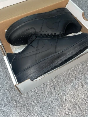 Nike Air Force 1  - Säljer ett par helsvarta Nike Air Force 1 sneakers i klassisk low-modell. Skorna är i läder med perforerad tå, svart sula och svarta skosnören. Ikonisk Nike Swoosh på sidorna och metallbricka på snörningen. Perfekta för en clean och stilren look.