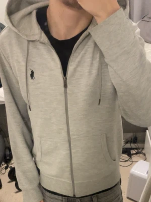 Ljusgrå hoodie från Ralph Lauren - Snygg ljusgrå hoodie med dragkedja från Ralph Lauren. Klassisk design med två fickor framtill, justerbar huva med dragsko och ikoniska loggan broderad på bröstet. Perfekt för en chill och stilren look.