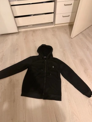 Svart hoodie från Polo Ralph Lauren - Säljer en svart hoodie med dragkedja från Polo Ralph Lauren. Klassisk design med huva och dragsnören, två fickor framtill och den ikoniska lilla loggan broderad på bröstet. Perfekt för en chill och stilren look.
