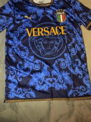 Blå  x Italia t-shirt - Unik blå t-shirt. T-shirten har ett detaljerat barockinspirerat mönster i blått och gult tryck, samt guldfärgade detaljer runt hals och nederkant. Tillverkad i polyester och har korta ärmar. Perfekt för dig som vill sticka ut.