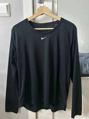 Svart långärmad topp från Nike - Svart långärmad topp från Nike med rund halsringning och klassisk vit Nike-logga framtill. Tillverkad i mjukt material som passar perfekt för träning eller en sportig look. Enkel och stilren design som är lätt att matcha med andra plagg.