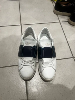 Valentino opens - Snygga vita sneakers från Valentino med bred mörkblå rem över vristen. Klassisk låg modell med vita skosnören och diskret logga på plösen. Tillverkade i slätt läder för en clean och exklusiv look. Väldigt bra skick! Givetvis äkta såklart!