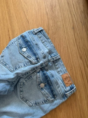 Ljusblå bootcut jeans från gina - Säljer ett par ljusblå jeans från gina  med bootcut passform och klassiska fickor med knappar bak. Jeansen har en snygg tvätt och är tillverkade i denim med skön stretch. Perfekta för dig som gillar en avslappnad och trendig look.