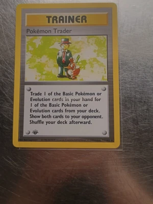 Pokémon Trader 1st Edition  - Säljer ett Pokémon Trainer-kort från Base Set. Kortet har gul ram, motiv av en man i kostym med en Pokéball och en Charmander. Texten är på engelska och baksidan har den klassiska blå designen med en Pokéball i mitten.