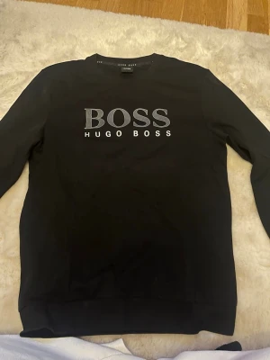 Svart sweatshirt från Hugo Boss - Svart sweatshirt från Hugo Boss med stort vitt BOSS-tryck och HUGO BOSS-text på bröstet. Klassisk rund halsringning och långa ärmar. Perfekt för en stilren och avslappnad look.