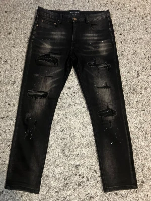 Supply & Demand NYC Svarta Jeans Distressed med Paint Splatter ”32w” - Säljer ett par svarta jeans från Supply & Demand med slitningar, hål och vita färgstänk för en edgy look. Jeansen har fem fickor, knappgylf och raka ben. Perfekta för dig som gillar streetstyle och vill sticka ut lite extra.