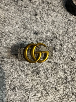 Gucci  bälte med guldspänne - Säljer ett Gucci bälte med klassiskt GG-mönster i beige och grå toner. Bältet har ett stort, rundat spänne i guld och är perfekt för att lyfta vilken outfit som helst. Ikonisk accessoar för dig som gillar lyxiga detaljer.
