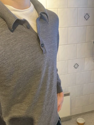 Grå henleytröja från Oscar Jacobson - Grå långärmad henleytröja från Oscar Jacobson med klassisk krage och tre knappar framtill. Tillverkad i mjukt material som ger en avslappnad och stilren look. Perfekt för dig som gillar enkel och tidlös design. Tröjan är märkt i XL men passar som S då den krympt i tvätten. Den har förvånansvärt kvar alla sina egenskaper så det är inget man märker av!