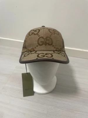 Beige keps med GG-mönster från Gucci - Säljer en beige keps från Gucci med det klassiska GG-monogrammet i brunt över hela kepsen. Kepsen har böjd skärm, justerbar rem bak och är tillverkad i canvasmaterial. Perfekt för dig som vill ha en exklusiv och trendig accessoar.