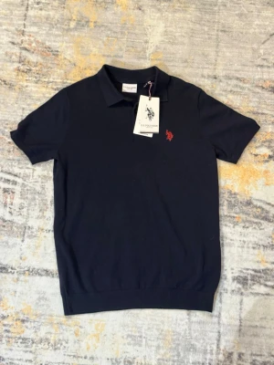 Marinblå pikétröja från U.S. Polo Assn. - Klassisk marinblå pikétröja från U.S. Polo Assn. med korta ärmar och röd broderad logga på bröstet. Tröjan har krage och knappar framtill, tillverkad i mjuk bomull som känns skön mot huden. Perfekt för en stilren och avslappnad look.