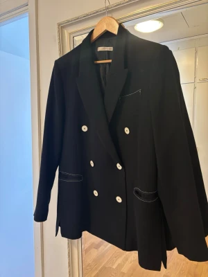 Svart dubbelknäppt kavaj med vita knappar - Stilren svart kavaj från Mango med dubbelknäppning och vita knappar. Kavajen har markerade slag, fickor med vit kontrastsöm och klassisk passform. Perfekt för dig som vill ha en snygg och trendig look. Den behöver strykas/steamas som ni kan se på sista bilden - så rätas det ut. 