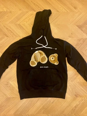 Svart hoodie  - Svart hoodie  med stor huva och vit snörning. På bröstet finns ett broderat motiv av en nallebjörn i beige och brunt. Hoodien är i relaxed passform. Perfekt för en streetwear-look.