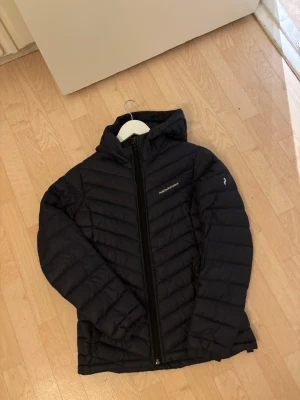 Svart dunjacka Peak Performance S - Snygg svart dunjacka från Peak Performance med huva och dragkedja framtill. Jackan har diskret logga på bröstet och är tillverkad i lätt, quiltat material som håller värmen. Perfekt för kalla vinterdagar och har en modern, smal passform.den är mörk blå men det svart ut 