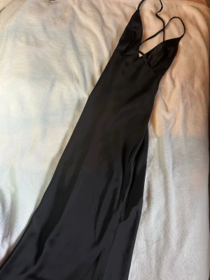 Svart lång satinklänning från Cider - Säljer en svart lång sidenklänning i satin. Klänningen har smala axelband som korsas i ryggen och en djup urringning framtill. 