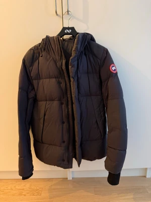 Svart dunjacka från Canada Goose - Säljer en svart dunjacka från Canada Goose med huva och klassisk patch på ärmen. Jackan har en puffig design, dragkedja och knappar framtill samt muddar vid ärmsluten. Perfekt för kalla vinterdagar och riktigt varm tack vare dunfyllningen. Över vänster armhåla så finns ett litet hål men det går lätt att lösa genom att sätta på en lagningslapp. Nypris 8995.