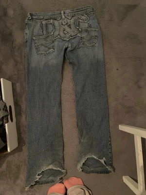 Blå D&G jeans med broderad logga - Säljer ett par blå jeans från D&G med stor broderad logga på bakfickorna. Jeansen har raka ben och slitna detaljer vid bensluten. Klassisk femficksmodell i denim med en cool vintagekänsla. Perfekta för dig som gillar streetwear och statement-plagg. Stora passar på M men lite baggy då.