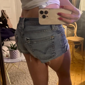 Blå jeansshorts från Levi's - Säljer ett par klassiska blå jeansshorts från Levi's med råa kanter och femficksmodell. Shortsen har låg midja och dragkedja fram till. Priset kan diskuteras då det finns flera hål i dom💓