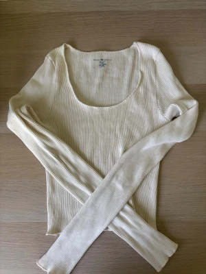 Ribbad beige croppad topp ifrån brandy melville - Toppen är bara avänd 1 gång. Toppen är i storlkeken one size men passar xs-s. Trendig ribbad topp i beige med croppad passform och lång ärm. Djup, rundad urringning framtill ger en snygg siluett. Perfekt att styla med höga jeans eller kjol för en clean look. 