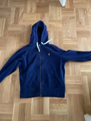 Marinblå hoodie från Polo Ralph Lauren - Snygg marinblå hoodie med dragkedja från Polo Ralph Lauren. Klassisk gul broderad logga på bröstet och vita dragsnören i huvan. Tillverkad i mjukt bomullsmaterial och har två fickor framtill. Perfekt för en avslappnad och stilren look.                                                            Priset kan diskuteras!