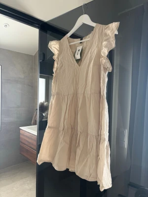 Beige volangklänning med v-ringning H&M  - Söt beige kortklänning med v-ringning och volangdetaljer vid ärmarna. Klänningen har en luftig passform och är tillverkad i ett lätt tyg, perfekt för varma dagar.