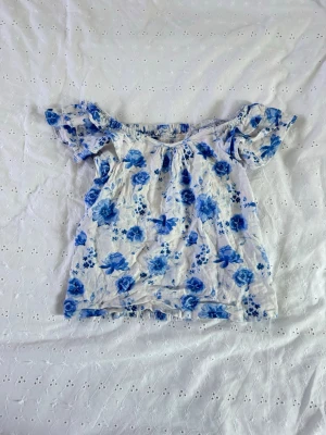 Blommig offshoulder topp i blått och vitt - Söt offshoulder topp med blått blommönster på vit botten. Puffiga korta ärmar och smockad rygg för skön passform. Perfekt till sommaren och lätt att matcha med jeans eller kjol.
