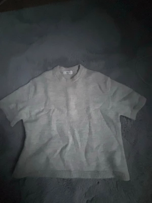 Ljusgrå stickad t-shirt Gina Tricot - Ljusgrå stickad t-shirt från Gina Tricot i ullblandning. Tröjan har korta ärmar, rund halsringning och ribbade kanter. Perfekt för lager-på-lager och enkel att matcha med olika outfits.