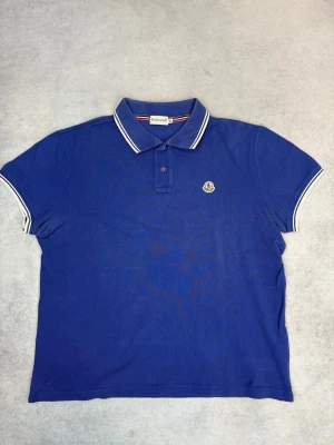 Blå pikétröja från Moncler - Klassisk blå pikétröja från Moncler med vit- och blårandig krage och ärmslut. Tröjan har korta ärmar, knappslå framtill och Moncler-logga på bröstet. Perfekt för en sportig och stilren look.