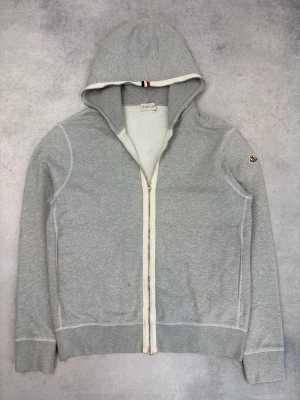 Ljusgrå hoodie från Moncler - Snygg ljusgrå hoodie från Moncler med dragkedja framtill och vit kontrastkant längs huvan och dragkedjan. Klassisk Moncler-logga på ärmen och ribbade muddar. Tillverkad i mjukt bomullsmaterial och har en sportig vibe med diskreta detaljer.