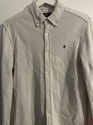 Randig skjorta från Morris - Snygg vit och grårandig skjorta från Morris med klassisk krage och broderad logga på bröstet. Skjortan har långa ärmar, nypris 1799 och mitt pris 299✨🌟, knappar framtill och är gjord i mjuk bomull. Perfekt för en clean och stilren look.