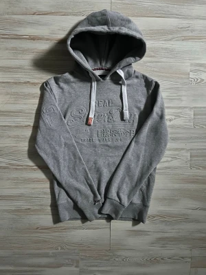 Grå hoodie från Superdry - Säljer en grå hoodie från Superdry med broderad logga och japansk text på bröstet🤗 Perfekt för dig som gillar en clean och sportig stil🤩 inga fel alls på hoodien. Hör av er vid funderingar🙌🏻