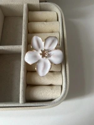 Vit blomformad ring med gulddetaljer - Säljer en superfin ring i form av en blomma med fem vita kronblad och små guldfärgade detaljer i mitten. Ringen har en guldfärgad stomme och är perfekt för dig som vill ha en söt och unik accessoar. Materialet är metall och emalj.